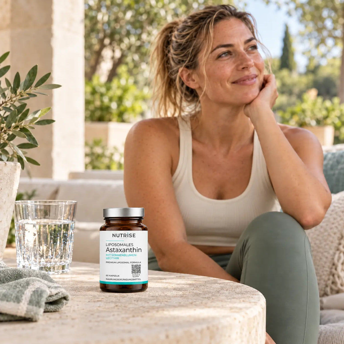 Nutrise Astaxanthin in realistischer Größe im Alltag: Frau auf einer Terrasse neben dem Produkt und einem Glas Wasser